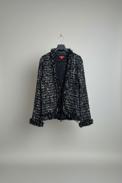 A black and white tweed open-front cardigan with frayed fringe details, hanging on a black hanger against  a studio background