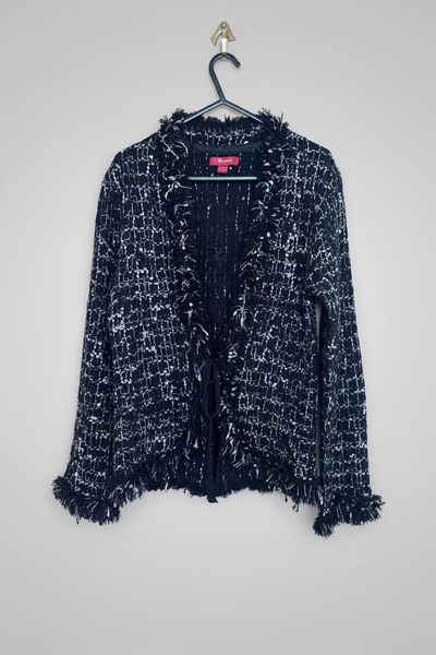 A black and white tweed open-front cardigan with fringed trim, hanging on a black hanger against a grey background