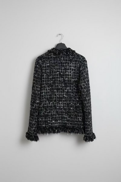 A black and white tweed cropped sweater with frayed fringe trim on the hem and cuffs, hanging on a black hanger against a studio background.
